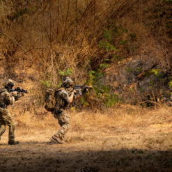 This image depicts three U.S. soldiers in full gear, marching in a line formation through a dry forest. They are dressed in camouflage clothing and are carrying rifles, indicating they are on patrol or in a combat situation. The background is filled with dry grass, trees, and rocks, adding to the realism of the scene. The warm color tone of the image enhances the overall atmosphere