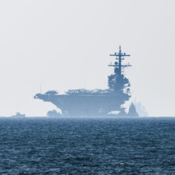 This image features a photo-realistic depiction of a dark blue aircraft carrier sailing on a light blue ocean, moving from left to right. Accompanying the carrier on its left side is a smaller boat. The sky above is a hazy gray, and the horizon line is visible in the distance, providing a sense of vastness to the scene. The image captures the majesty and power of naval operations.