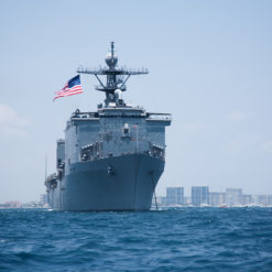 This image showcases a large, gray U.S. Navy ship sailing on the deep blue ocean, with small waves visible around it. The ship is equipped with a prominent radar tower and is flying an American flag on its mast, symbolizing its origin. The ship is moving towards the right side of the image. In the background, a city skyline under a clear blue sky can be seen, providing a stark contrast to the vastness of the ocean.