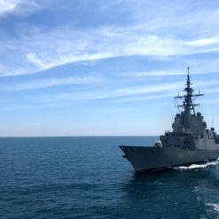 This image captures a large, gray naval ship sailing on the deep blue ocean. The ship is equipped with a towering structure, housing multiple antennas and radar dishes, indicative of its advanced communication and surveillance capabilities. The ship is seen from the side, showcasing its impressive size and structure. Above, the sky is a clear blue with a few white clouds scattered across. The image encapsulates the might and presence of naval power on the vast ocean.