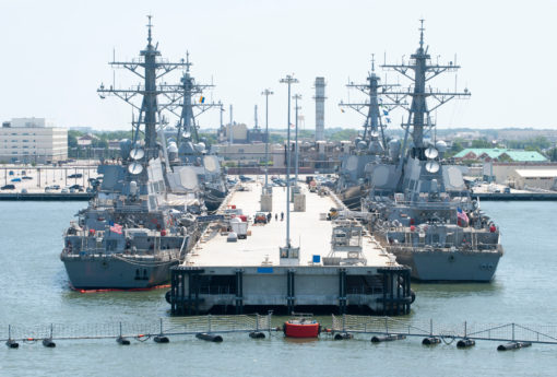 This image features two large, grey navy ships docked at a concrete pier. The ships are multi-leveled and equipped with various antennas, signifying their advanced capabilities. On the pier, several smaller boats and vehicles can be seen, indicating activity. A red and white striped barrier marks the end of the pier. The calm water in the foreground is dotted with several buoys. In the background, a city skyline under a clear blue sky provides a contrasting backdrop to the naval scene.