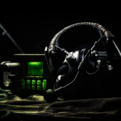 A photo-realistic image of a black headset with a microphone, resting on a camouflage fabric. The headset is connected to a black device with a green screen and buttons, resembling a radio or communication device. The image has a dark and moody tone, emphasizing the seriousness of military communication equipment.