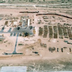 This image provides an aerial view of a military construction site located in a desert-like area. The site is characterized by sandy terrain and is populated with various structures and vehicles. On the right side of the image, there are several rows of rectangular structures, possibly storage containers. The left side features cranes and other construction vehicles. A long, straight road bisects the site. The overall color scheme is a dusty brown, with sparse greenery visible in the background.