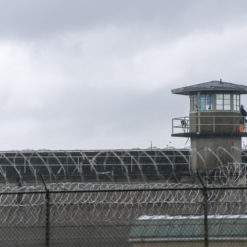 A photo-realistic image of a prison watchtower, a concrete structure with a blue roof and windows on all sides. In the foreground, there’s a chain-link fence topped with barbed wire. The sky is overcast, contributing to the overall bleak mood of the image, indicative of the harsh realities of prison environments.