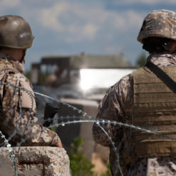 This image depicts two U.S. soldiers standing behind a concrete barrier topped with barbed wire. Both soldiers are dressed in camouflage uniforms and helmets, with the soldier on the left wearing a green helmet and the soldier on the right wearing a desert camouflage helmet. The backdrop is a clear blue sky with clouds, and a military vehicle can be seen in the distance. The setting appears to be a war zone or military base.