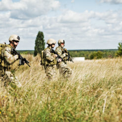 This image portrays three U.S. soldiers in full camouflage gear, walking through a tall, dry field. Each soldier is equipped with a rifle and a helmet, signifying readiness for action. The field they traverse is expansive, with trees visible in the distance. Above them, the sky is a clear blue, dotted with white clouds. To respect their privacy, the faces of the soldiers are intentionally blurred.