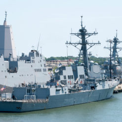This image depicts a group of U.S. Navy ships docked at Norfolk Harbor. The ships, painted in standard naval grey, are multi-leveled with various towers and structures. Each ship proudly flies the American flag. The harbor water is a calm light blue, with smaller boats also docked nearby. The backdrop of the image features the city skyline of Norfolk, Virginia, complete with buildings and trees.