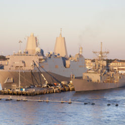 A tranquil harbor scene during golden hour showcases two prominent U.S. naval ships moored side by side. The ship to the left is a modern gray destroyer with distinct angular pyramid-like structures (known as stealthy Advanced Gun Systems) and a flatter deck surface. To its right is a slightly older navy vessel with a more traditional silhouette, featuring various antennas and a central superstructure. Both vessels are adorned with U.S. flags. The waterfront has several structures and service vehicles attending to the ships, and the calm waters in the foreground have a series of buoys. The backdrop is illuminated by the soft light of the setting sun, casting a warm hue over everything.