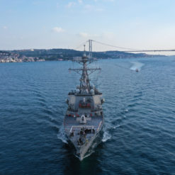 Aerial view of a large, gray U.S. naval ship sailing on blue water towards a city skyline with a large suspension bridge. The ship features a prominent tower in the center and is surrounded by small waves. The sky above is blue with a few clouds.
