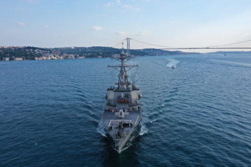 Aerial view of a large, gray U.S. naval ship sailing on blue water towards a city skyline with a large suspension bridge. The ship features a prominent tower in the center and is surrounded by small waves. The sky above is blue with a few clouds.