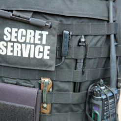 A photo-realistic image of a person’s torso wearing a black tactical vest with ‘SECRET SERVICE’ written on it in white letters. The vest is equipped with multiple pockets and pouches, and a radio is attached to it. The person is also wearing a black belt with a holster and a knife sheath attached to it. The background is blurred, focusing attention on the detailed equipment of the Secret Service personnel.