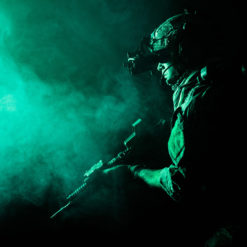 A side-profile view of a U.S. military soldier, illuminated by a dim, eerie green light. The soldier wears a helmet equipped with night-vision goggles, positioned down over his eyes. He is dressed in tactical gear and holds a rifle close to his body, ready for action. The surrounding atmosphere is filled with a thick, swirling mist or smoke, creating a sense of tension and anticipation. The soldier's expression is focused and determined, highlighting the intensity of night-time operations and the readiness of the U.S. military personnel.