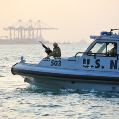 This image captures a U.S. Navy boat, number 503, navigating choppy waters. The boat is a small, motorized vessel painted in a standard naval grey. A military personnel, fully geared, stands on the bow of the boat, holding a gun. In the hazy background, a large cargo ship with cranes can be seen. The water around the boat is disturbed, indicating the boat’s swift movement.