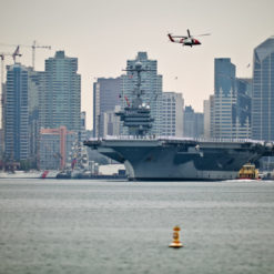 Foreground depicts calm harbor waters with a buoy floating nearby. Dominating the midground is a massive US aircraft carrier anchored close to the shore. Above it, a helicopter is in flight, moving towards the cityscape. The background is a sprawling urban skyline with a mixture of tall residential buildings, office towers, and ongoing construction sites with cranes. The city's architectural features range from glass-clad modern structures to more traditional buildings. The atmosphere appears overcast, casting a soft light over the entire scene, blending the military might of the carrier with the everyday life of the city.