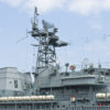 Detailed view of a naval warship's upper structures. The ship's grey superstructure is dominated by a towering mast loaded with various communication and radar equipment, including circular and spherical antennas. Ladders and platforms are visible around the mast, allowing personnel access. On the ship's deck, multiple covered lifeboats are aligned in a row. The background is a light blue sky with some cloud cover, emphasizing the ship's readiness and technological prowess against a calm backdrop.