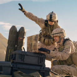 The image portrays a group of United States Marines in a desert setting, engaged in an operation. The Marines, dressed in desert camouflage uniforms and helmets, are positioned around a large weapon that appears to be a missile launcher. One Marine stands and points towards an unseen target, while two others kneel next to the weapon, ready for action. The backdrop is a vast desert landscape with distant mountains under a clear, blue sky.