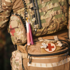 Close-up of a U.S. military soldier's torso, highlighting various equipment details. The soldier wears a camouflaged uniform, and around the waist is a tactical belt with pouches. Prominently displayed on one of the pouches is a white cross on a red background, symbolizing medical aid. Above this pouch are shotgun shells secured in place. An antenna with a pinkish protective cover extends upward, suggesting communication equipment. A hint of a rifle is seen on the left, with its sling draped across the chest. The image emphasizes the dual role of combat and medical assistance provided by some soldiers.