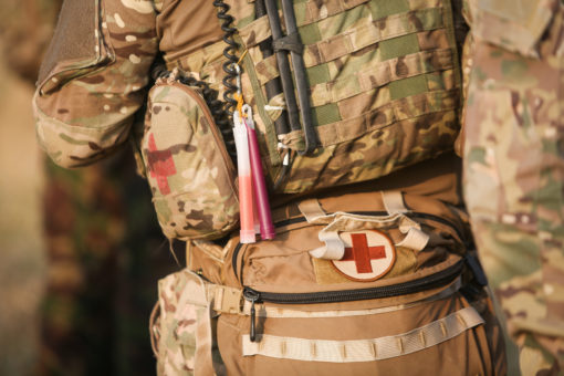 Close-up of a U.S. military soldier's torso, highlighting various equipment details. The soldier wears a camouflaged uniform, and around the waist is a tactical belt with pouches. Prominently displayed on one of the pouches is a white cross on a red background, symbolizing medical aid. Above this pouch are shotgun shells secured in place. An antenna with a pinkish protective cover extends upward, suggesting communication equipment. A hint of a rifle is seen on the left, with its sling draped across the chest. The image emphasizes the dual role of combat and medical assistance provided by some soldiers.