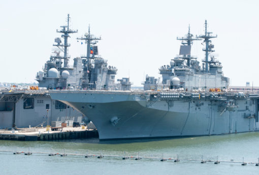 Two large U.S. naval warships are moored side-by-side at a port, their massive gray hulls and towering superstructures with multiple radar domes, antennas, and communication masts dominate the view. Both ships have flight decks at the rear, suggesting they are equipped to handle helicopters or aircraft. In the foreground, calm waters reflect the ships' silhouettes, and a series of buoys floats in a line parallel to the vessels. The backdrop provides a clear sky, hinting at a serene day at the naval base.