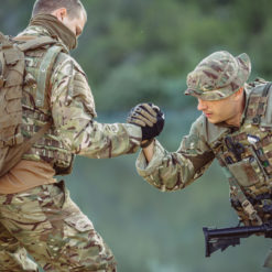Two soldiers in camouflage uniforms in a forested backdrop. The soldier on the left, carrying a large backpack, reaches out with gloved hands to assist the soldier on the right, who is in a semi-crouched position with a rifle slung in front. The interaction suggests camaraderie and teamwork in a field setting.
