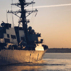 The image presents a breathtaking view of a U.S. Navy destroyer ship at sunset. The ship, characterized by its gray color, radar tower, and multiple guns and missile launchers, is stationed in calm waters. The sun sets behind the ship, painting the sky in shades of orange and pink. The serene water reflects the captivating hues of the sunset, with the ship silhouetted against this vibrant backdrop.
