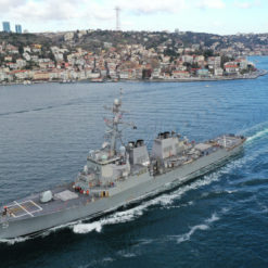 The USS Donald Cook (DDG 75), a U.S. Naval Destroyer, sails in deep blue waters. Recognizable by its gray hull and the number "75" painted near the bow, the ship is equipped with advanced radar systems atop and a helipad at the rear. In the background, a densely built coastal city ascends on the hills, showcasing diverse architecture and a few tall structures. The waterway provides a distinct separation between the ship and the city, highlighting the interplay of military presence and civilian landscapes.