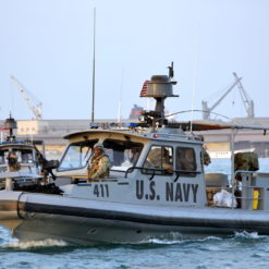 This image features a US Navy patrol boat, identifiable by the number 411 on its side, stationed in a bustling harbor. The boat, painted in a standard naval gray, is small in size and equipped with a radar dome on top and a compact cabin. The backdrop of the image reveals a busy harbor scene, complete with cranes and other boats. Captured during the day, the sky above is a clear blue.
