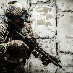 Close-up image of a soldier in full tactical gear against a backdrop of a weathered brick wall. The soldier wears a camouflaged uniform and a helmet equipped with night-vision goggles. Their face is partly obscured by the goggles and other gear, but focus and determination can be sensed in their posture. The soldier holds a modern assault rifle with both hands, ready for action. Various pouches, cables, and attachments are evident on the uniform, indicating preparedness for diverse combat scenarios. The worn-out bricks in the background have patches of old plaster, providing a stark contrast to the soldier's modern equipment. The overall tone is dramatic, emphasizing the intensity and readiness of the moment.