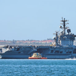 A massive U.S. aircraft carrier, with the number '66' prominently displayed on its superstructure, is moored at a harbor. The carrier, with its distinctive flight deck, is accompanied by a smaller tugboat sailing nearby. The waters are calm, and in the distance, a coastal city landscape is faintly visible under a clear sky.