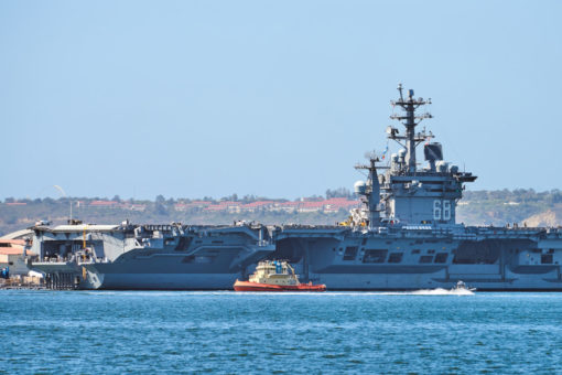 A massive U.S. aircraft carrier, with the number '66' prominently displayed on its superstructure, is moored at a harbor. The carrier, with its distinctive flight deck, is accompanied by a smaller tugboat sailing nearby. The waters are calm, and in the distance, a coastal city landscape is faintly visible under a clear sky.