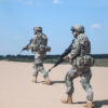 Two soldiers in desert camouflage gear traverse a sandy field under a clear blue sky. The soldier on the left carries a rifle, while the one on the right, slightly ahead, shoulders a machine gun. Both soldiers wear helmets with face shields, moving in unison in the same direction.