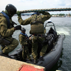 Daytime scene of three US Navy SEALs, equipped with helmets, body armor, and weapons, preparing to board a black inflatable Zodiac-type boat on a concrete pier. A large rope barrier is visible on the pier. The backdrop features a serene body of water with a distant city skyline.