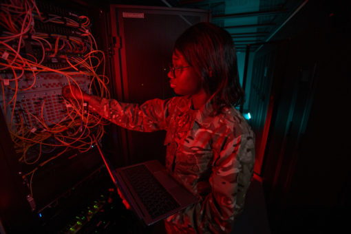 A focused military technician, wearing camouflage uniform and glasses, is working within a dimly lit server room. Illuminated by the soft glow of red lights from the server racks, she attentively examines a panel of tangled colorful cables while holding an open laptop in her other hand. The racks contain various network equipment, with blinking lights indicating active connections. The atmosphere is one of concentration and expertise as the technician troubleshoots or configures the system. The narrow space between server racks further emphasizes the close-quarters nature of the technical work. The image portrays the blend of military discipline with modern technology expertise.