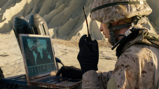 A United States Marine Corps soldier, dressed in a helmet and camouflage uniform, sits in a desert setting surrounded by sandbags. The soldier is engaged with a laptop computer that displays a map. With one hand, the soldier points at the screen, indicating a location on the map.