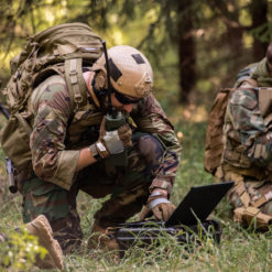 The image depicts a group of U.S. soldiers in a forest setting. The soldiers are dressed in camouflage clothing and are carrying backpacks, indicating a field operation. One soldier is kneeling on the ground, engrossed in something on a laptop. The other soldiers are standing around the kneeling soldier, their attention focused on the laptop screen. The backdrop of the image is a dense forest, providing a natural camouflage for the soldiers.