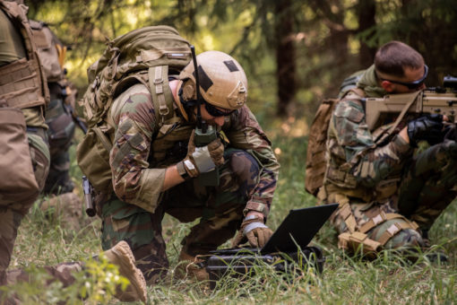 The image depicts a group of U.S. soldiers in a forest setting. The soldiers are dressed in camouflage clothing and are carrying backpacks, indicating a field operation. One soldier is kneeling on the ground, engrossed in something on a laptop. The other soldiers are standing around the kneeling soldier, their attention focused on the laptop screen. The backdrop of the image is a dense forest, providing a natural camouflage for the soldiers.
