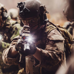 In a scene dense with tension, a group of U.S. special operations soldiers are captured in the midst of an operation. The central figure, wearing glasses, is intently aiming his rifle fitted with a tactical flashlight, its beam piercing through the haze. He's clad in a camouflage uniform, a helmet with mounted equipment, and a face mask, giving a sense of both protection and anonymity. To his left, another soldier can be seen similarly equipped, his own weapon's light joining the ensemble of focused beams. Surrounding them are blurred outlines of fellow operatives, emphasizing the rapid movement and urgency of the moment. The overall atmosphere is one of coordination, readiness, and intense concentration amidst the challenges of their mission.