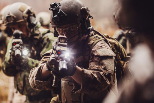 In a scene dense with tension, a group of U.S. special operations soldiers are captured in the midst of an operation. The central figure, wearing glasses, is intently aiming his rifle fitted with a tactical flashlight, its beam piercing through the haze. He's clad in a camouflage uniform, a helmet with mounted equipment, and a face mask, giving a sense of both protection and anonymity. To his left, another soldier can be seen similarly equipped, his own weapon's light joining the ensemble of focused beams. Surrounding them are blurred outlines of fellow operatives, emphasizing the rapid movement and urgency of the moment. The overall atmosphere is one of coordination, readiness, and intense concentration amidst the challenges of their mission.