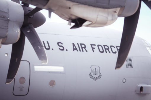 This image features a U.S. Air Force aircraft in flight, captured from a low angle. The aircraft, painted white, proudly displays “U.S. Air Force” in blue lettering on its side. A shield emblem with a white eagle against a blue background is also visible on the aircraft’s body. The aircraft is equipped with four propellers, two of which are visible in this shot. The backdrop of the image is a cloudy sky, emphasizing the aircraft’s airborne status.