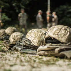 The image presents a row of military helmets and vests arranged on the ground. The helmets, adorned with hanging straps, and the vests both sport a camouflage pattern. They rest on a surface of grass and dirt. In the background, blurred figures of soldiers can be seen standing and walking around. The image employs a shallow depth of field, drawing focus to the helmets and vests in the foreground.