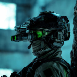 The image showcases an individual equipped with a night vision helmet, complete with a green visor and a gas mask. The helmet is intricately detailed with multiple attachments and wires. The person is dressed in a camouflage jacket and positioned in front of a tree trunk. The background, intentionally blurred, hints at the silhouette of a building with a window. The entire image is bathed in a blue-green color tone, emphasizing the night-time setting.