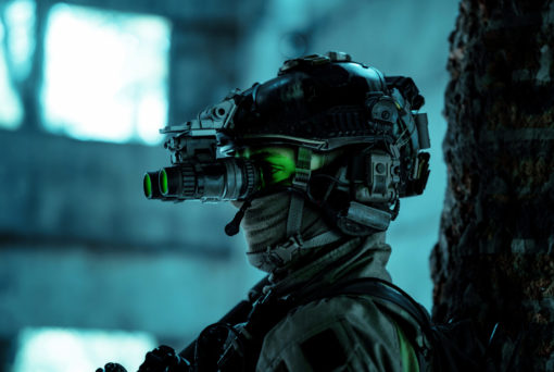 The image showcases an individual equipped with a night vision helmet, complete with a green visor and a gas mask. The helmet is intricately detailed with multiple attachments and wires. The person is dressed in a camouflage jacket and positioned in front of a tree trunk. The background, intentionally blurred, hints at the silhouette of a building with a window. The entire image is bathed in a blue-green color tone, emphasizing the night-time setting.
