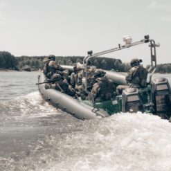 This is a black and white photo of a group of soldiers in a military inflatable boat, moving through the water and creating a wake. The soldiers are equipped with helmets and weapons. The boat is fitted with a motor and a spotlight. The background reveals a body of water with trees lining the shore.