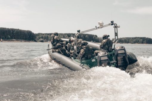 This is a black and white photo of a group of soldiers in a military inflatable boat, moving through the water and creating a wake. The soldiers are equipped with helmets and weapons. The boat is fitted with a motor and a spotlight. The background reveals a body of water with trees lining the shore.