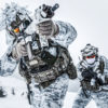 Two soldiers dressed in winter camouflage gear are poised in a combat stance. They are equipped with firearms and are wearing white suits with grey and black accents for camouflage. Their helmets are fitted with goggles and face masks. The backdrop is a snowy landscape with distant trees, and the image has a blurred effect around the edges, focusing attention on the soldiers.