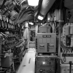 This is a black and white photo of the interior of a submarine, showing a narrow corridor filled with pipes, valves, and gauges on the walls and ceiling. Two control panels are visible on the right side of the photo, one labeled “Pump Room” and the other “Control Room”. The floor is covered in a metal grating. The photo appears to be taken from the perspective of someone standing in the corridor looking towards the control panels.