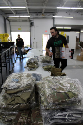 This is a photo of a warehouse with a long table in the center, covered with stacks of folded clothing, mostly in shades of green and brown. There are two people in the photo, the person in the foreground is wearing a black t-shirt with “Police” written on it in green letters. The background consists of shelves filled with boxes and other items.