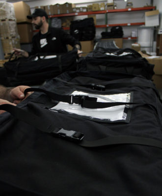 This is a photo of a pile of sturdy black bags stacked in a warehouse. Each bag has a white label on it with a barcode and some text.