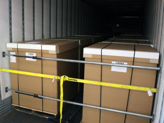 This is a photo of a stack of brown cardboard boxes on a pallet inside a shipping container. The boxes have white labels on them that read “SSI” and “CONTROL”. The pallet is secured with a yellow strap. The background is the inside of a shipping container, with the door open and light coming in from outside.
