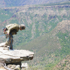 A person in a camouflage outfit and helmet is skillfully rappelling down a rocky cliff. The individual is secured with a rope and is leaning forward, indicating the action of descent. The cliff, composed of rocks and boulders, forms part of a mountainous landscape adorned with trees and shrubs. The person is positioned on a rock ledge, ready to navigate the challenging terrain.
